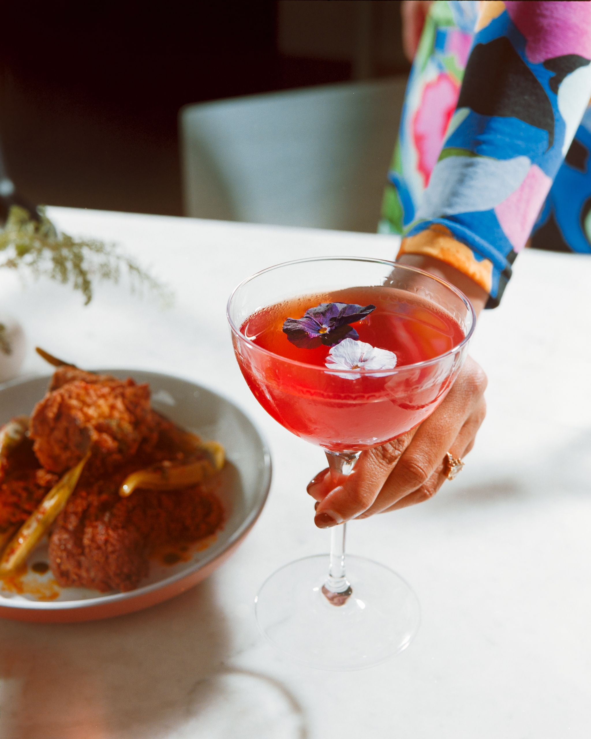 A person wearing a colorful floral dress holds a coupe glass filled with a bright pink cocktail garnished with edible flowers. A plate with fried food is blurred in the background.
