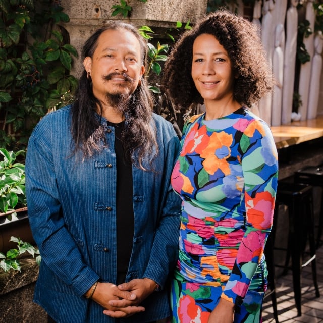 A portrait of a man with long hair and a mustache wearing a blue textured shirt, standing next to a woman with curly hair wearing a vibrant multi-colored floral print dress, outdoors surrounded by greenery.