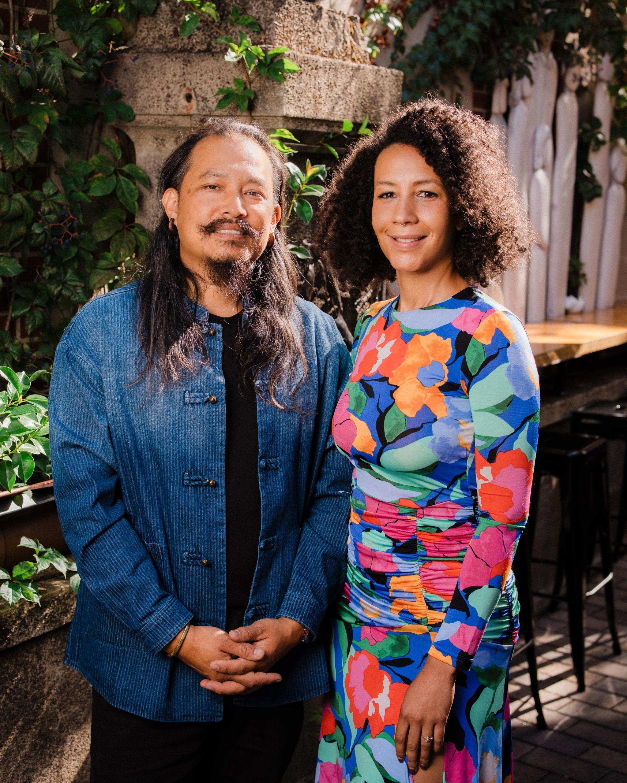 A portrait of a man with long hair and a mustache wearing a blue textured shirt, standing next to a woman with curly hair wearing a vibrant multi-colored floral print dress, outdoors surrounded by greenery.
