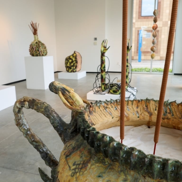 An art gallery exhibition of abstract ceramic sculptures in a white room with a concrete floor. In the foreground is a large, boat-shaped sculpture with thick, dark edges and tall reeds rising from a bed of sand.
