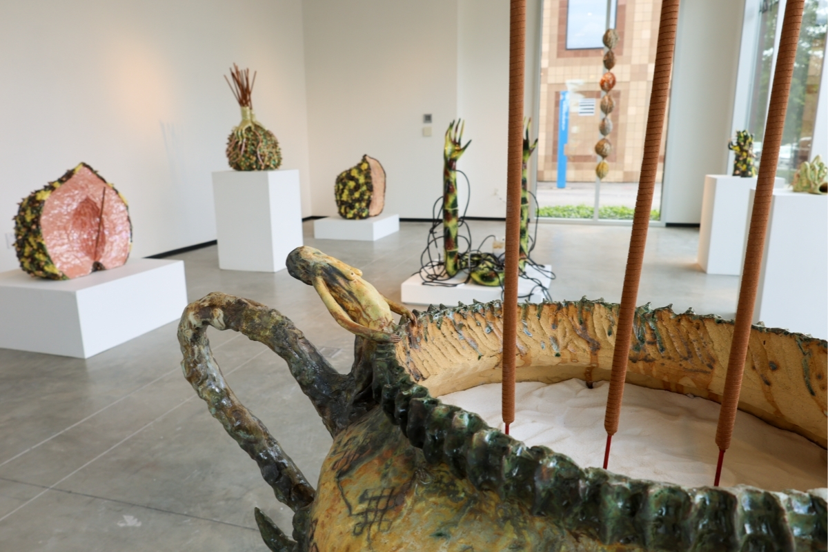 An art gallery exhibition of abstract ceramic sculptures in a white room with a concrete floor. In the foreground is a large, boat-shaped sculpture with thick, dark edges and tall reeds rising from a bed of sand.