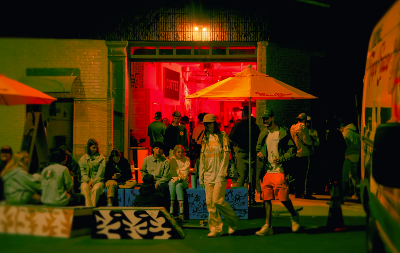 A vibrant, low-light photo of a crowd gathered outside a building entrance illuminated with intense red and orange light, featuring people sitting on wooden steps and standing under large umbrellas at night.
