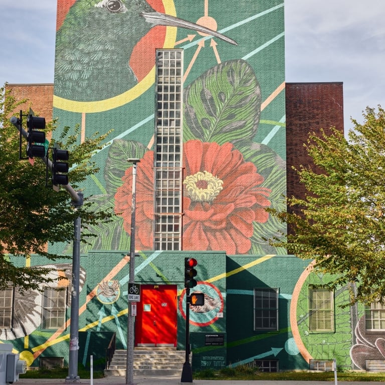 A massive mural on the exterior of a brick building depicting a large green hummingbird and a red flower with geometric background patterns. A tall window runs through the center of the artwork, and a red doorway is visible at the base.