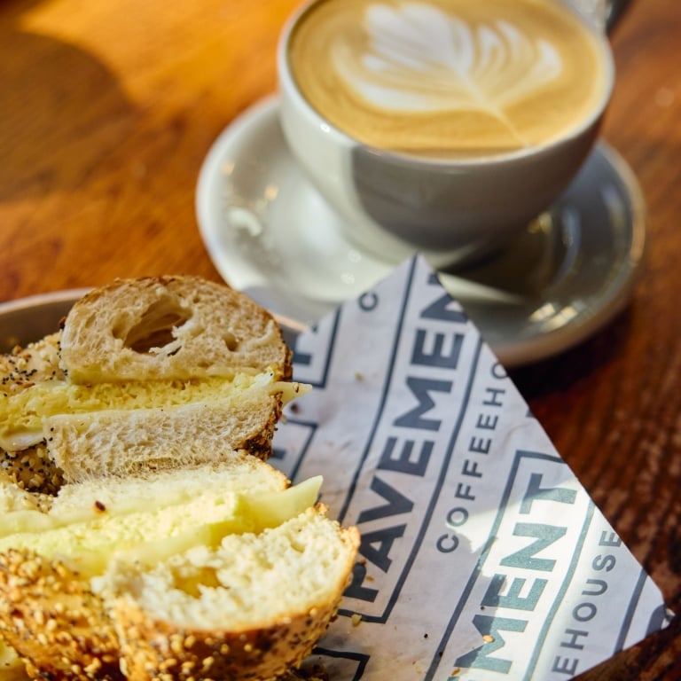 A close-up of a bagel sandwich with egg and cheese, sliced in half and resting on a branded paper napkin, next to a white mug of latte with beautiful leaf art on the wooden table.
