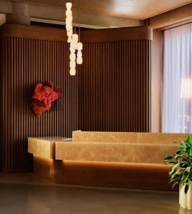 The Atlas front desk in travertine stone beneath stacked pendant lights, with fluted wood paneling and a sculptural red wall artwork.