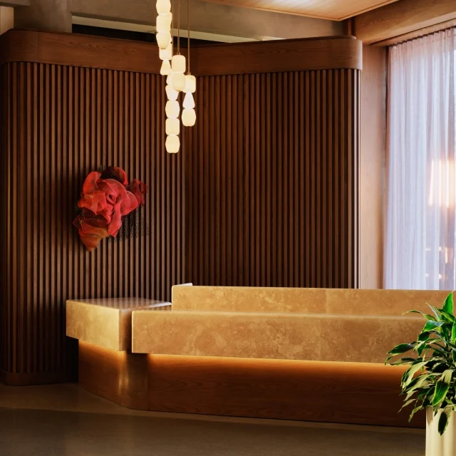 The Atlas front desk in travertine stone beneath stacked pendant lights, with fluted wood paneling and a sculptural red wall artwork.