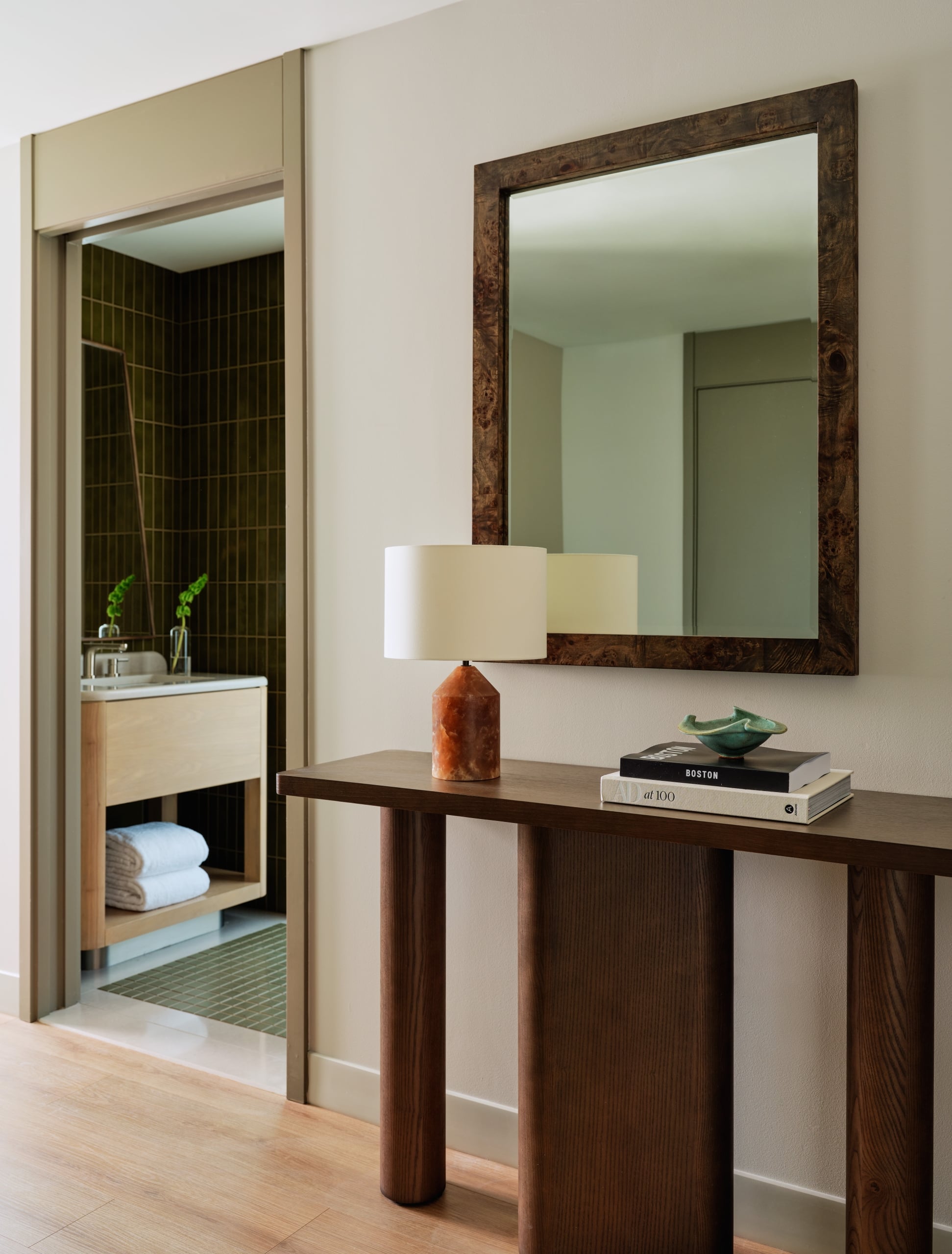 Room entryway with a dark wood console, burl mirror, ceramic lamp, and a glimpse into a green-tiled bathroom beyond.