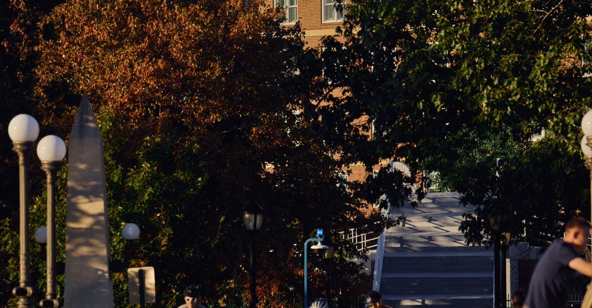 Cyclists and pedestrians cross the Weeks Bridge in Cambridge at golden hour, with autumn trees and a historic brick building beyond.