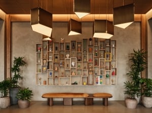 The Atlas hotel lobby with a curio shelf wall of eclectic art objects, a leather bench, tropical plants, and sculptural pendant lighting above.