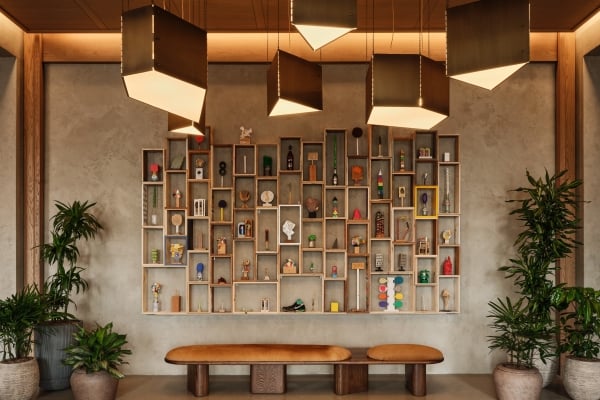 The Atlas hotel lobby with a curio shelf wall of eclectic art objects, a leather bench, tropical plants, and sculptural pendant lighting above.