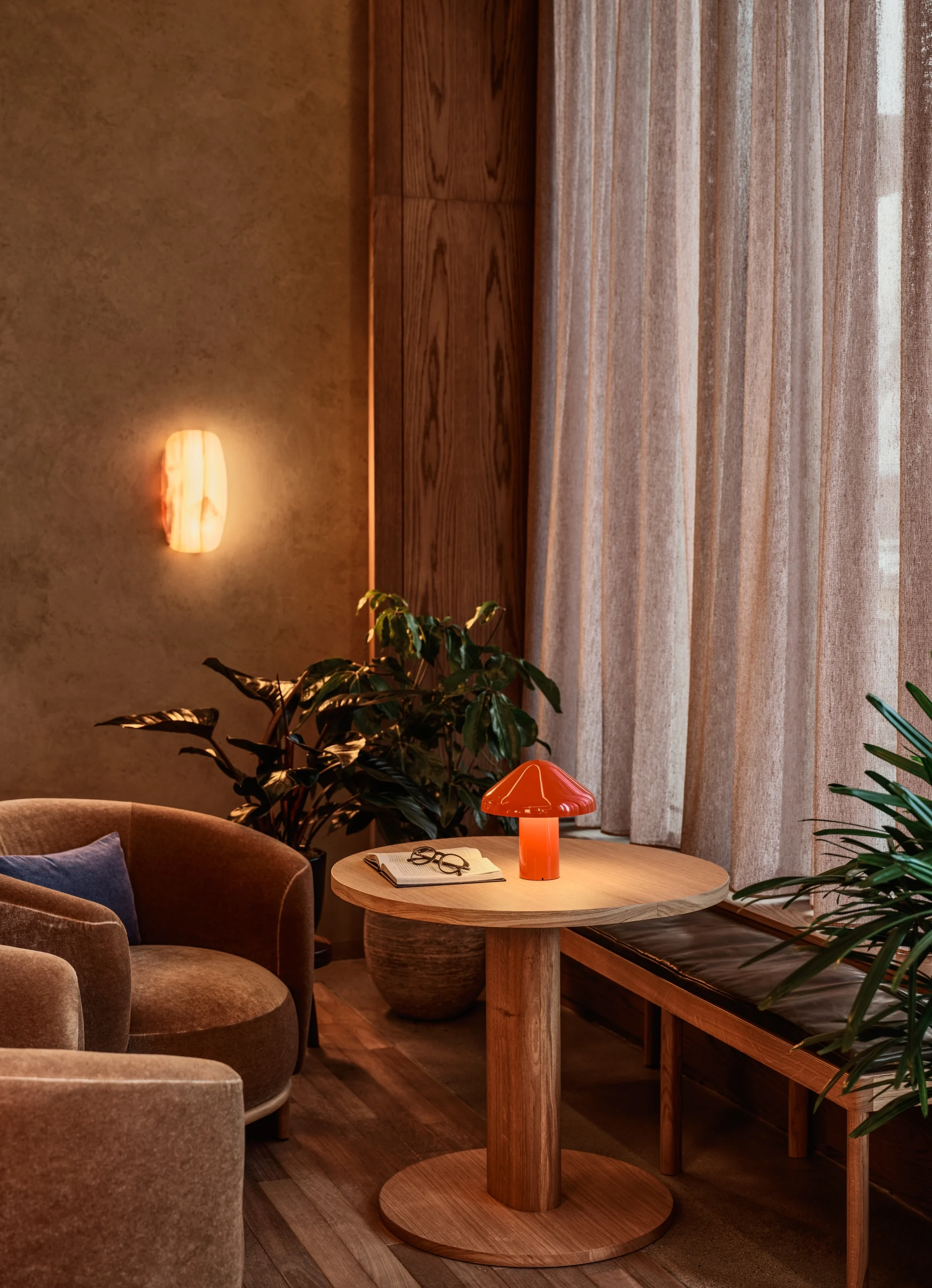 A warm lounge nook with armchairs, a round wood side table, an orange mushroom lamp, and a glowing alabaster wall sconce.