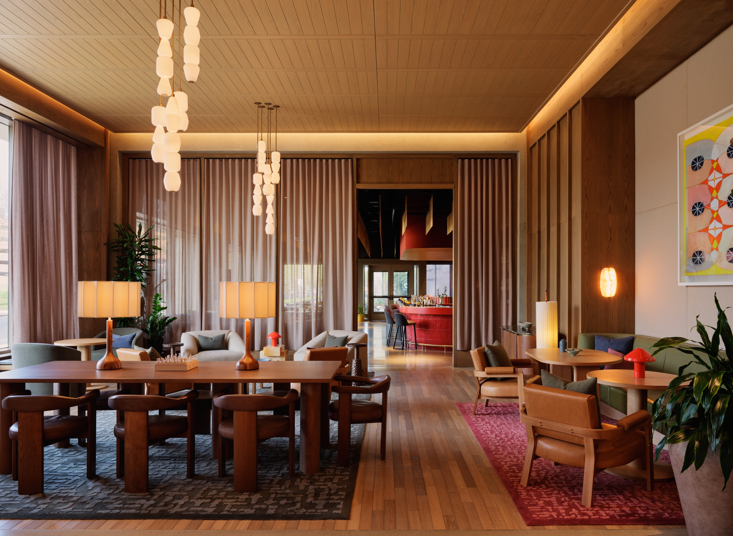 The Atlas hotel lobby lounge with warm wood paneling, pendant lighting, leather seating, and a glimpse of the red bar in the background.