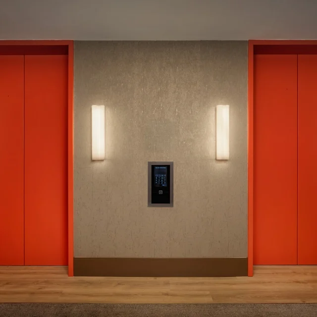 Two bright orange elevator doors framed by red walls in a modern lobby, with a central wall featuring a keypad panel and vertical wall lights.