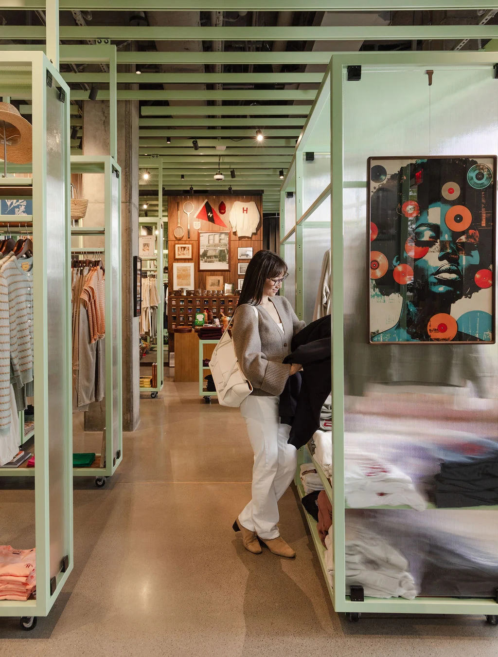 Woman in a gray cardigan and white pants browses folded clothes on a rack inside a store with mint-green shelving nearby.