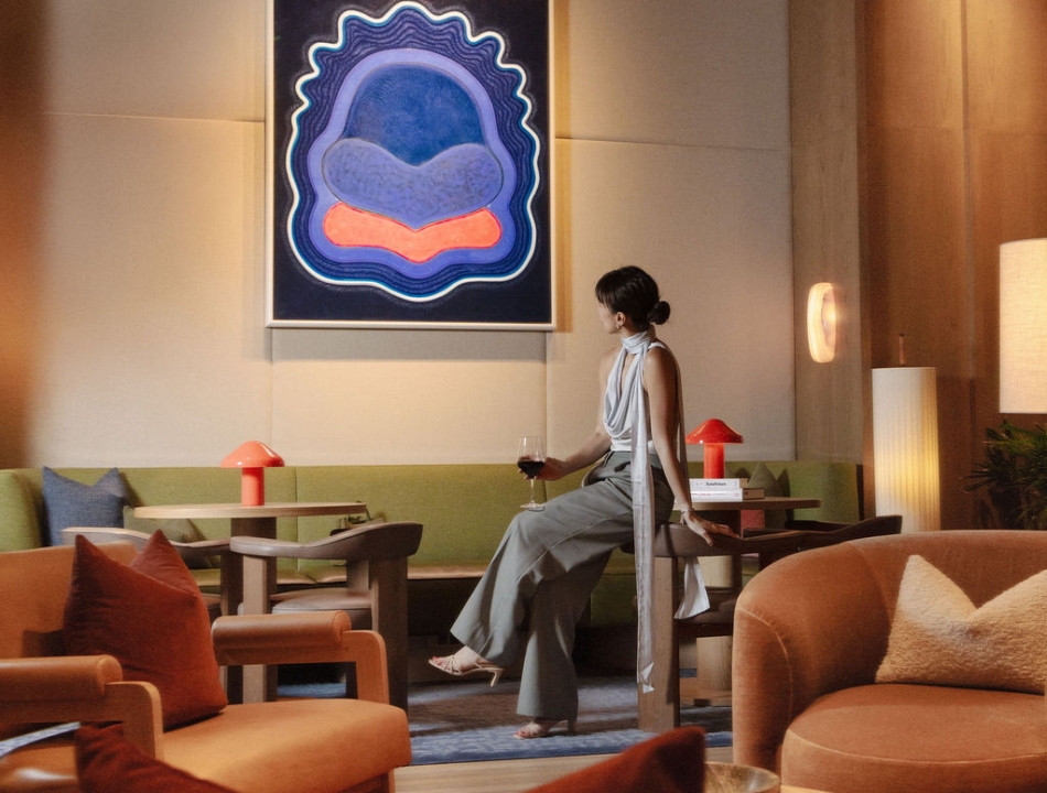 Woman in a grey sleeveless dress sits on a bench in a stylish hotel lounge, holding a glass of red wine, with a blue abstract painting on the wall behind.