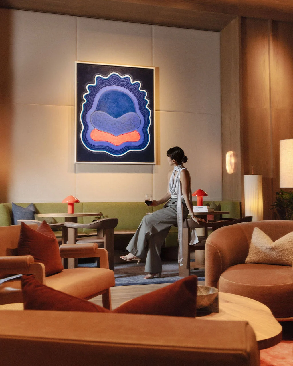 Woman in a grey sleeveless dress sits on a bench in a stylish hotel lounge, holding a glass of red wine, with a blue abstract painting on the wall behind.