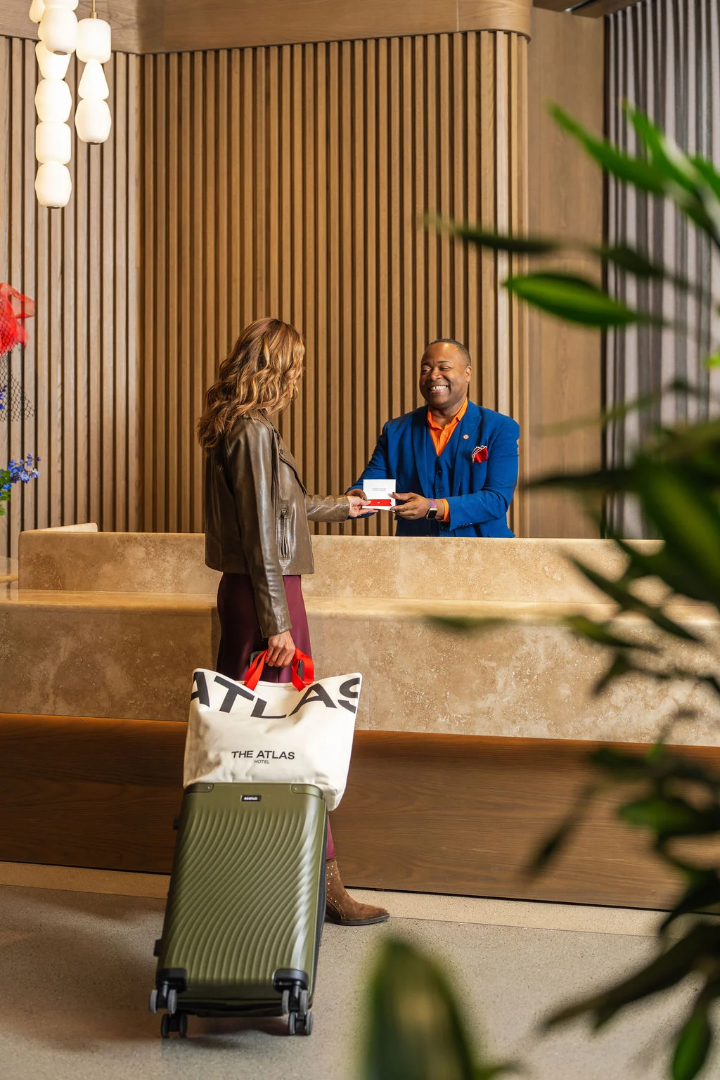 Hotel lobby check-in: guest with green suitcase and Atlas tote receives key from a smiling receptionist in a blue blazer.
