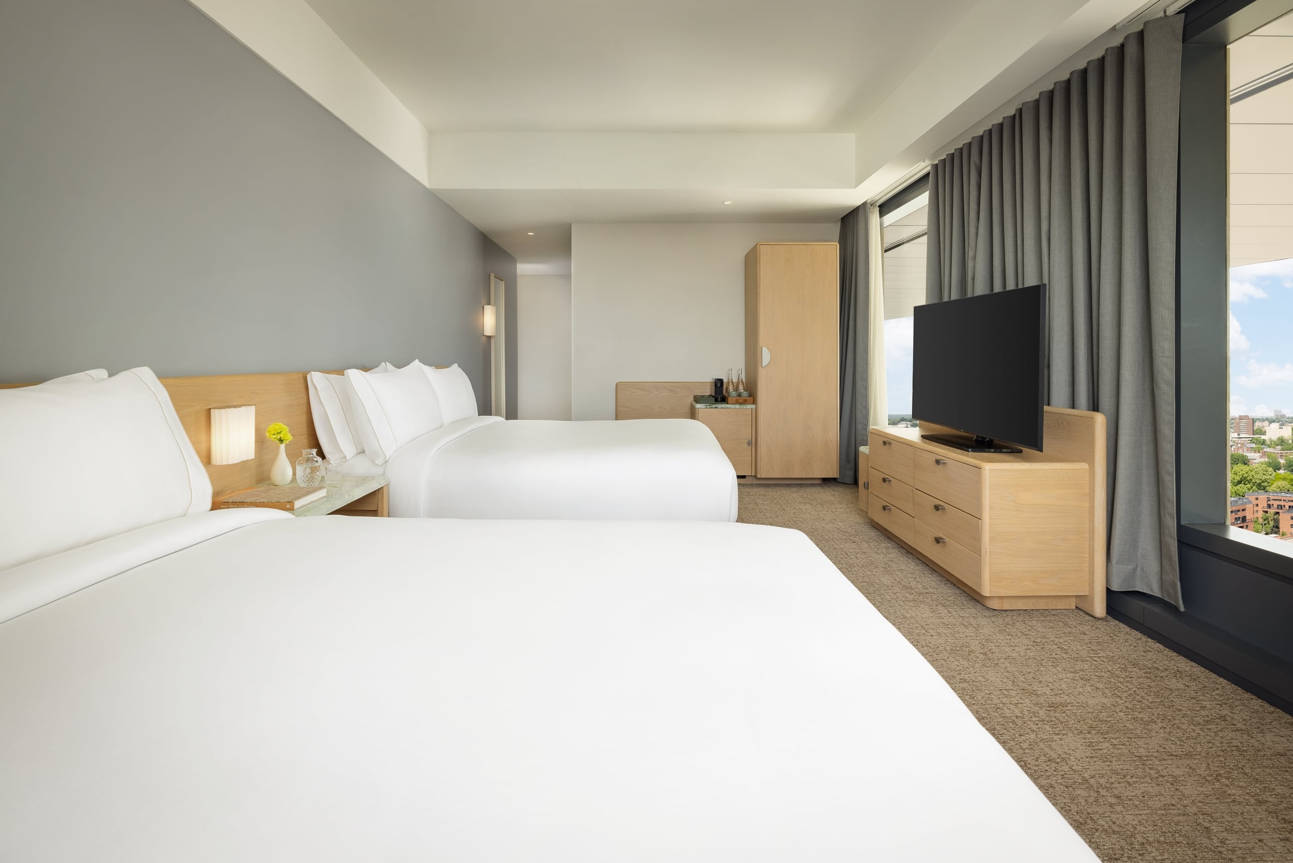 Hotel room with two white beds, nightstands, and a flat-screen TV facing the beds. Large window with gray curtains on the right.