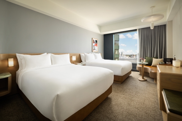Hotel room with two white-duvet beds, wooden headboards, bedside lamps, and a seating area by a large window overlooking the city skyline.