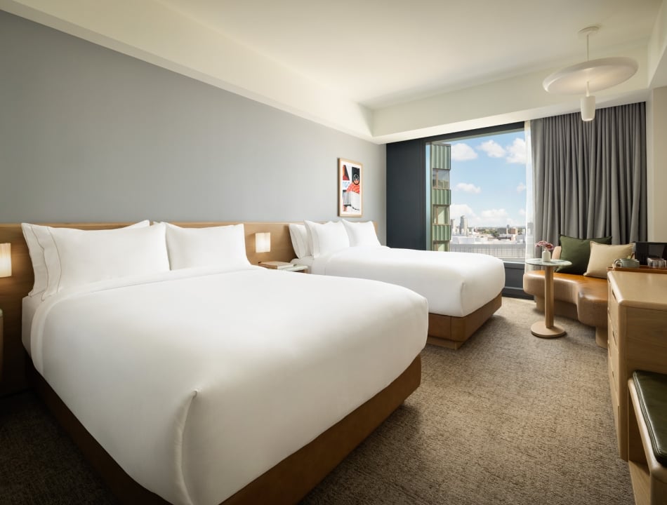 Hotel room with two white-duvet beds, wooden headboards, bedside lamps, and a seating area by a large window overlooking the city skyline.