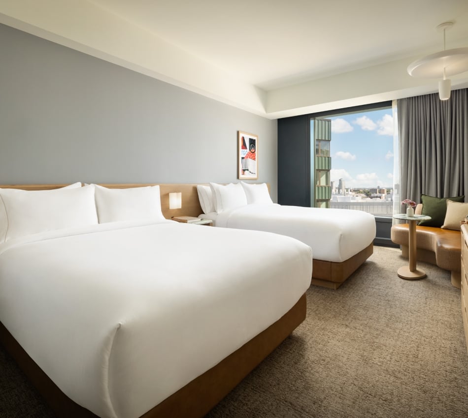 Hotel room with two white-duvet beds, wooden headboards, bedside lamps, and a seating area by a large window overlooking the city skyline.