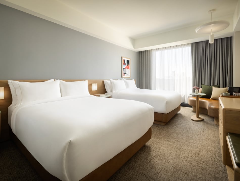 Hotel room with two neatly made white beds and a wooden headboard, by a large window with sheer curtains and a seating area nearby.