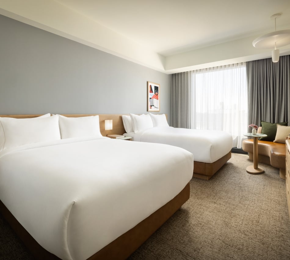 Hotel room with two neatly made white beds and a wooden headboard, by a large window with sheer curtains and a seating area nearby.