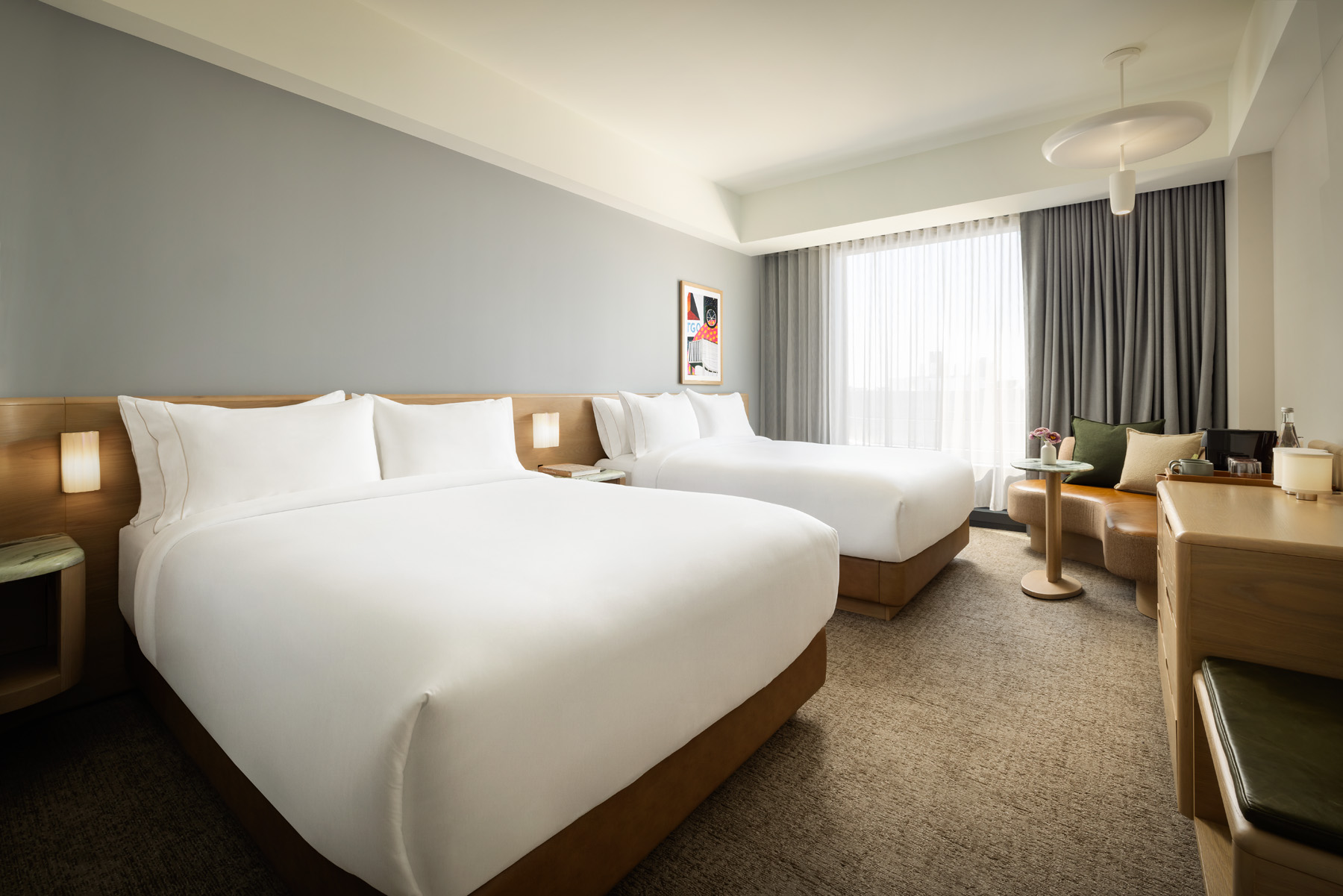 Hotel room with two neatly made white beds and a wooden headboard, by a large window with sheer curtains and a seating area nearby.