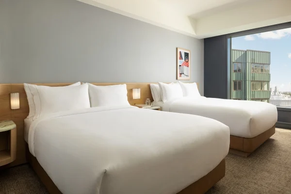Hotel room with two large white beds, wooden headboard, bedside lamp, and a windowside view of a green building and cityscape.