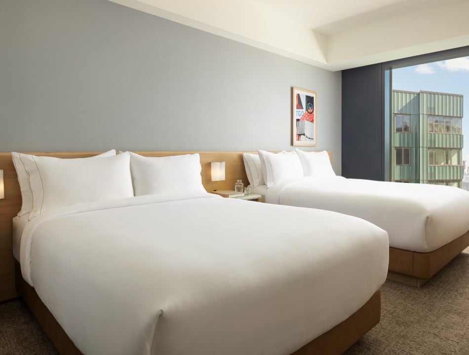 Hotel room with two large white beds, wooden headboard, bedside lamp, and a windowside view of a green building and cityscape.