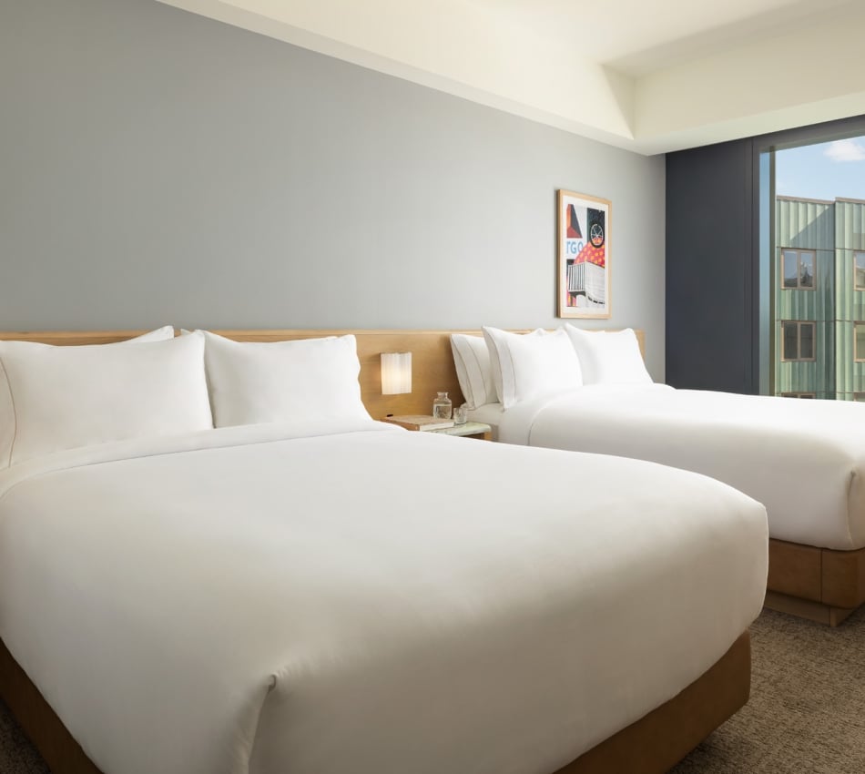 Hotel room with two large white beds, wooden headboard, bedside lamp, and a windowside view of a green building and cityscape.