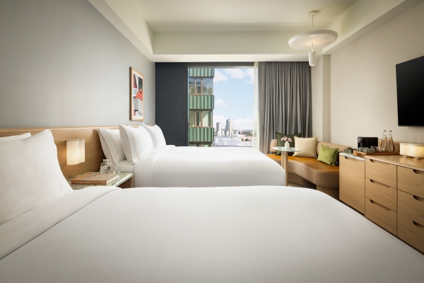 Hotel room with two white beds, nightstands, and a seating area by a large window overlooking city buildings.