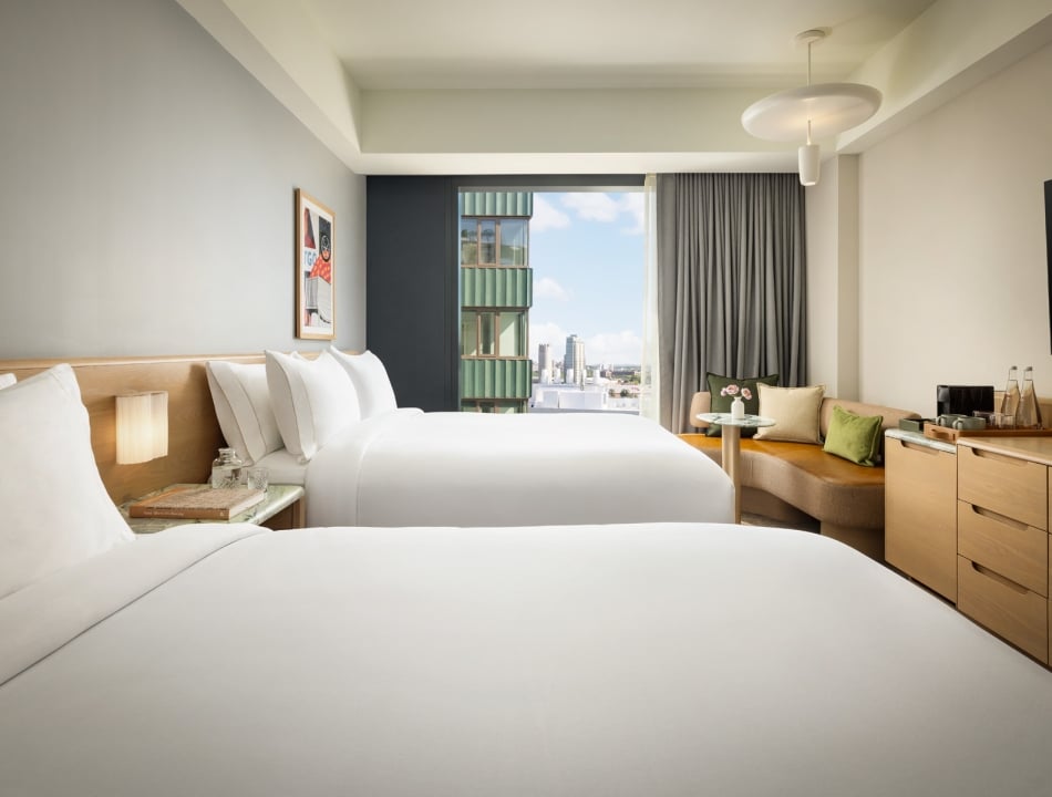 Hotel room with two white beds, nightstands, and a seating area by a large window overlooking city buildings.
