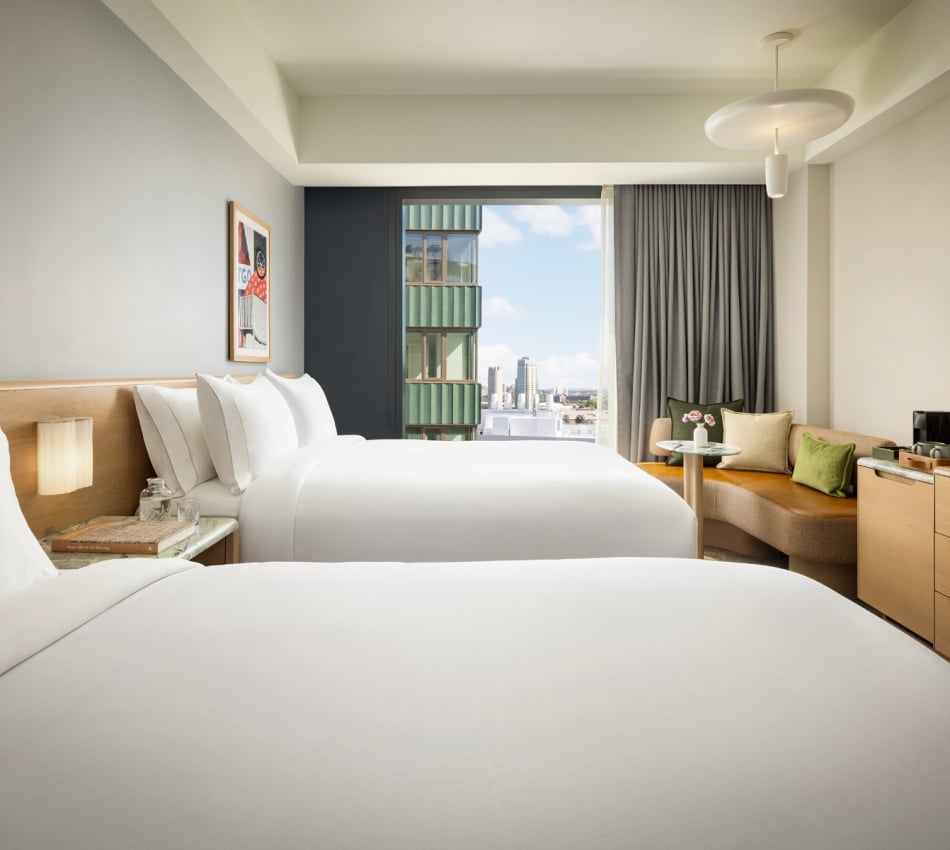 Hotel room with two white beds, nightstands, and a seating area by a large window overlooking city buildings.