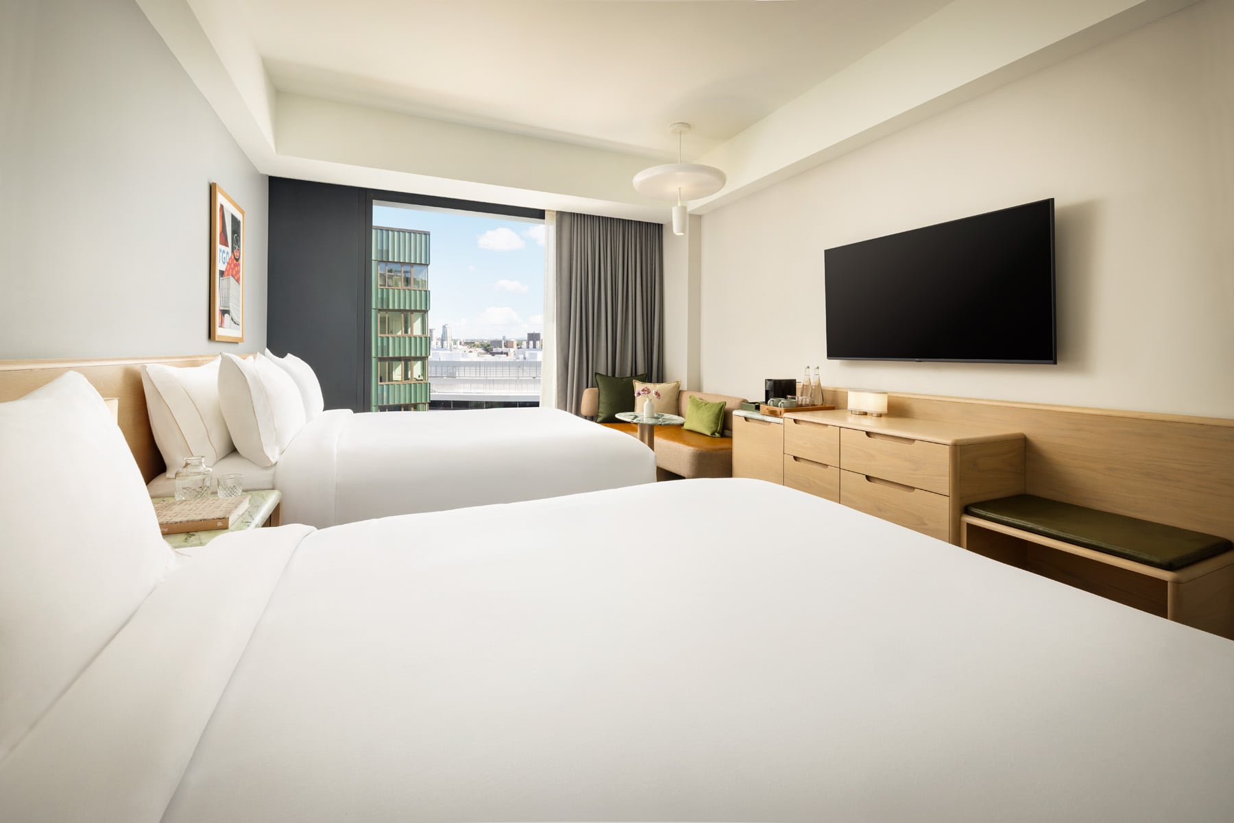 Hotel room with two white beds, wooden headboard, and a large window with a city view and gray curtains.