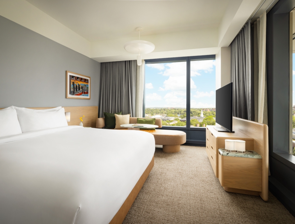 Modern hotel room with a large bed, seating area by a window, and a TV on a wooden dresser; city view outside the window.