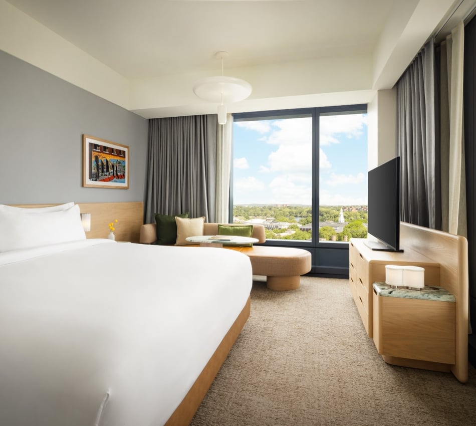 Modern hotel room with a large bed, seating area by a window, and a TV on a wooden dresser; city view outside the window.