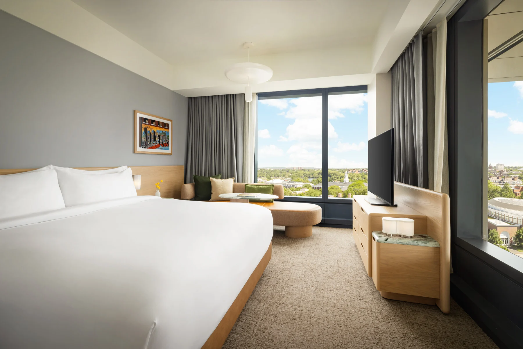 Modern hotel room with a large bed, seating area by a window, and a TV on a wooden dresser; city view outside the window.