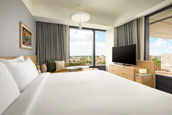 Hotel room interior with white bedding, gray walls, large windows, and a flat-screen TV on a wooden dresser overlooking the city skyline.