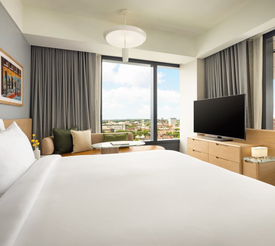 Hotel room interior with white bedding, gray walls, large windows, and a flat-screen TV on a wooden dresser overlooking the city skyline.