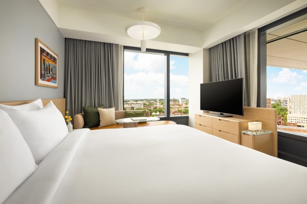 Bright hotel room with a large bed in the foreground, seating by a window, and a flat-screen TV on a wooden dresser overlooking the city skyline at dusk or day.