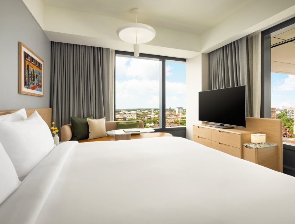 Bright hotel room with a large bed in the foreground, seating by a window, and a flat-screen TV on a wooden dresser overlooking the city skyline at dusk or day.