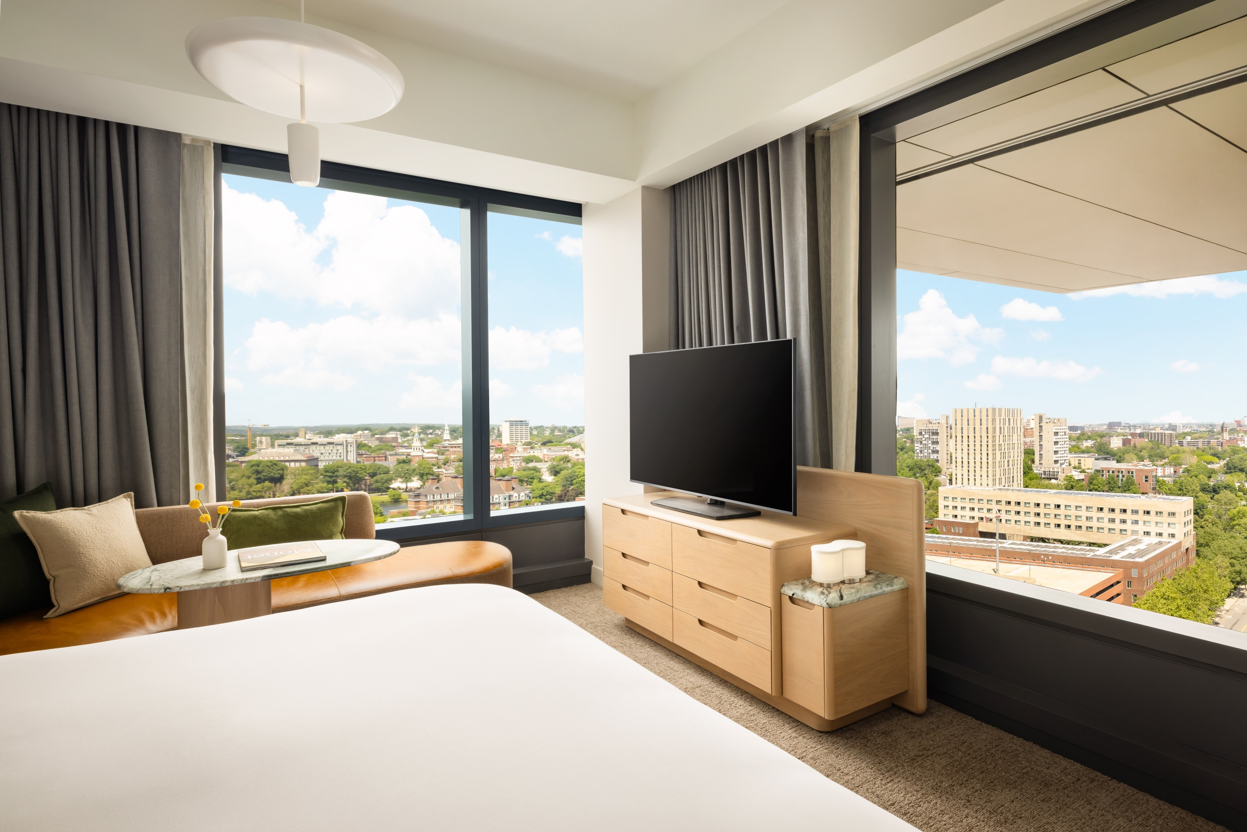 Hotel suite interior with large windows showing a city skyline, TV on a wooden dresser, and a beige sofa with cushions in the foreground.