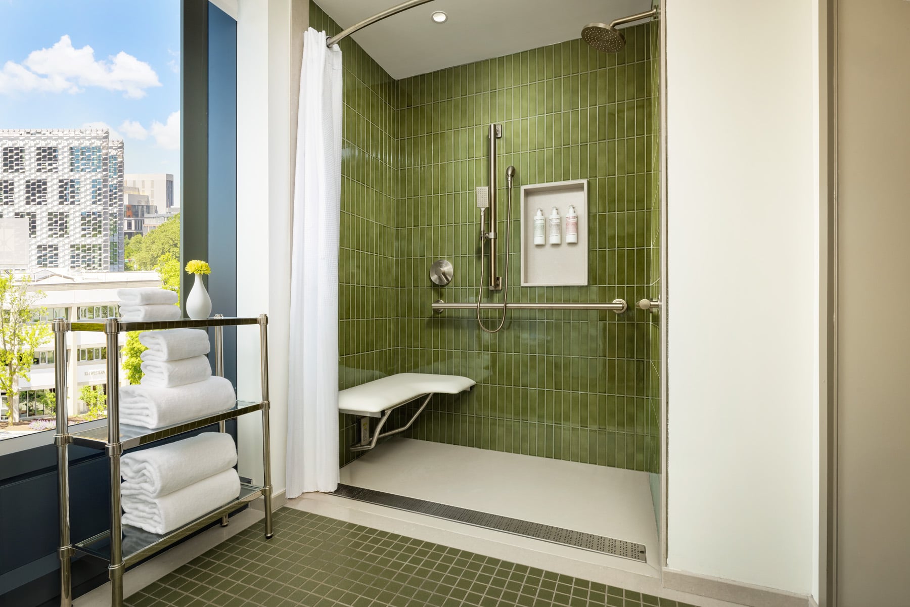 Hotel bathroom with green-tiled walk-in shower and built-in shower controls; glass exterior view with city buildings and blue sky outside.