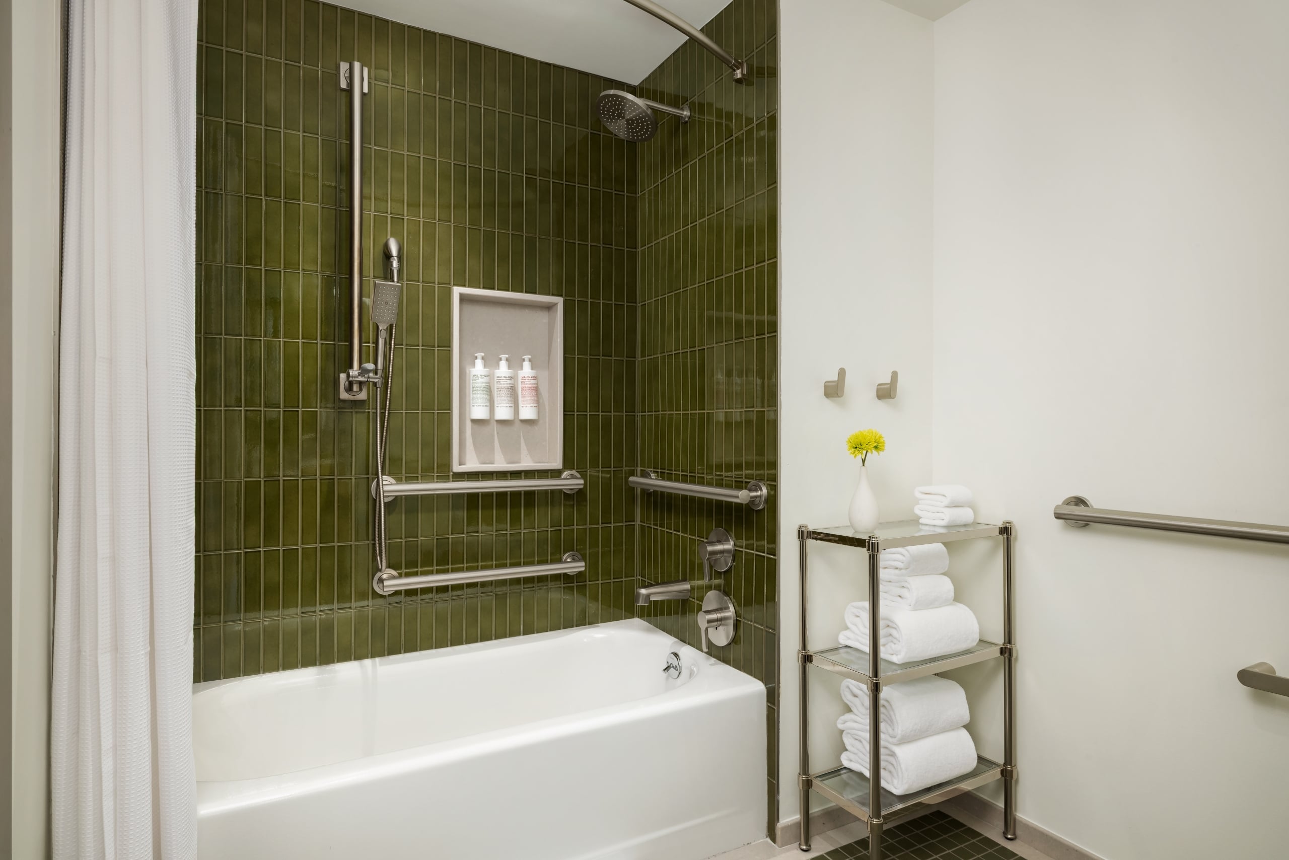 Modern bathroom shower area with dark green tile walls, rain showerhead, handheld shower, and grab bars, plus a built‑in niche with toiletries.