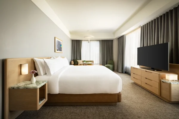 King-size bed with white bedding, bedside tables, and a seating area by large windows with heavy curtains in a modern hotel room.