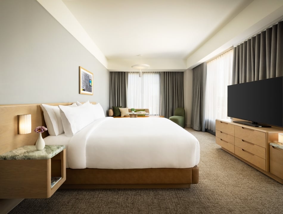 King-size bed with white bedding, bedside tables, and a seating area by large windows with heavy curtains in a modern hotel room.