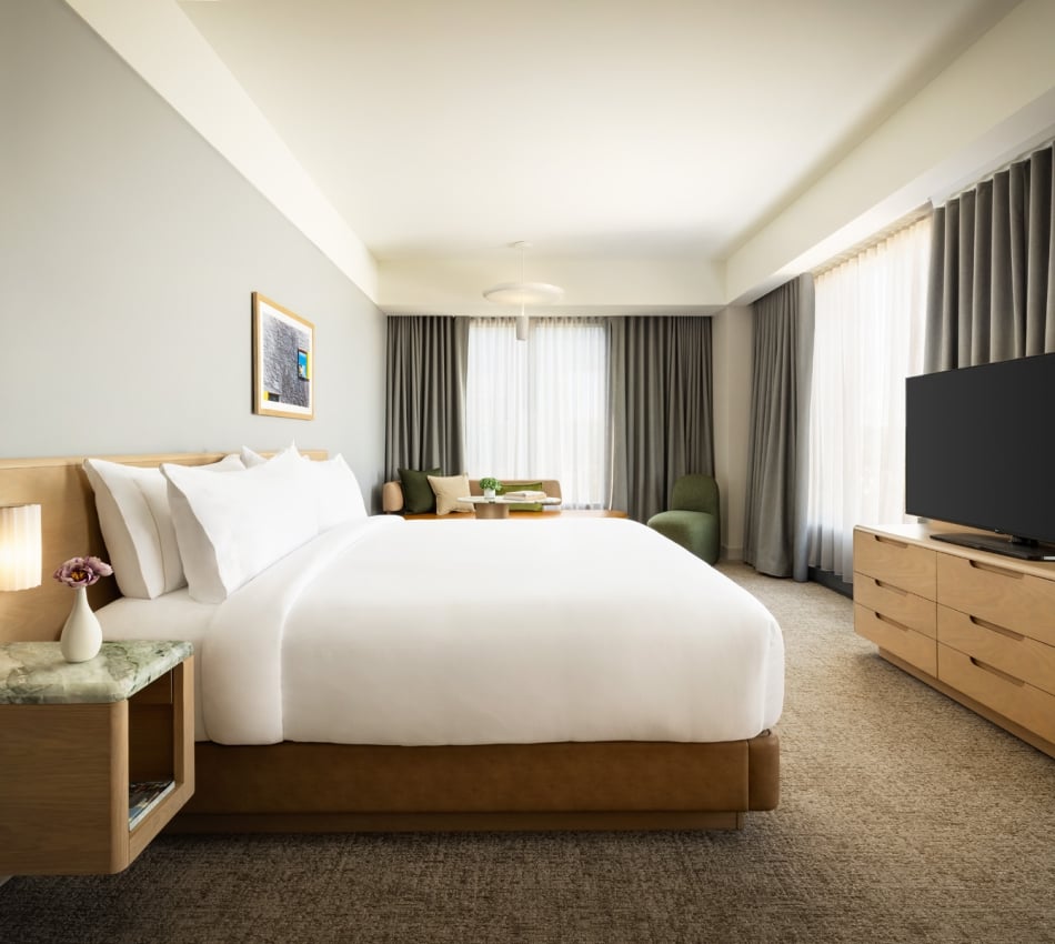 King-size bed with white bedding, bedside tables, and a seating area by large windows with heavy curtains in a modern hotel room.