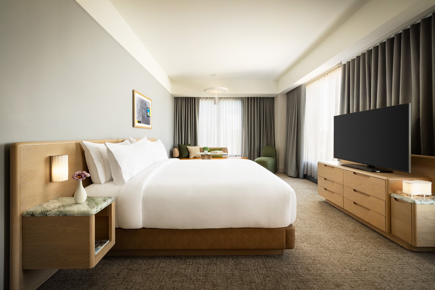 King-size bed with white bedding, bedside tables, and a seating area by large windows with heavy curtains in a modern hotel room.
