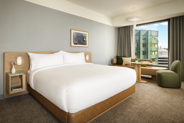 King-size bed with white bedding and a light wood headboard, a small bedside table with a vase of flowers, and a framed picture on a gray wall in a bright hotel room.